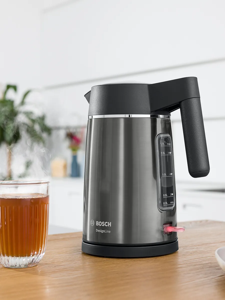 გრაფიტისფერი Bosch DesignLine ელექტრო ჩაიდანი დგას ხის ზედაპირზე ცხელი ჩაის ჭიქის გვერდით.
