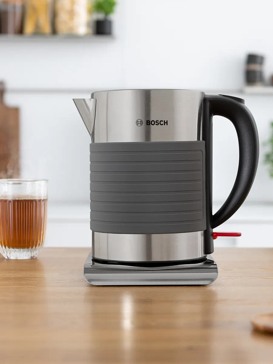 უჟანგავი ფოლადის Bosch-ის ელექტრო ჩაიდანი ნაცრისფერი სილიკონის სახელურით და ძირით, ხის ზედაპირზე, მის გვერდით კი დგას ჭიქა ჩაი.