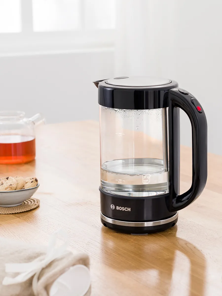 ალტერნატიული ტექსტი: შავი და გამჭვირვალე Bosch-ის ელექტრო ჩაიდანი (1.7 ლ, 2400 ვტ) ხის მაგიდაზე, ფონზე მინის ჩაიდანი და ნამცხვრები.