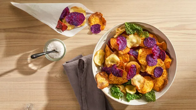 Una ciotola e un cono di colorate chips di verdura – incluse varietà viola, arancioni, bianche e verdi – con un barattolo di sale e un tovagliolo grigio su un tavolo di legno.