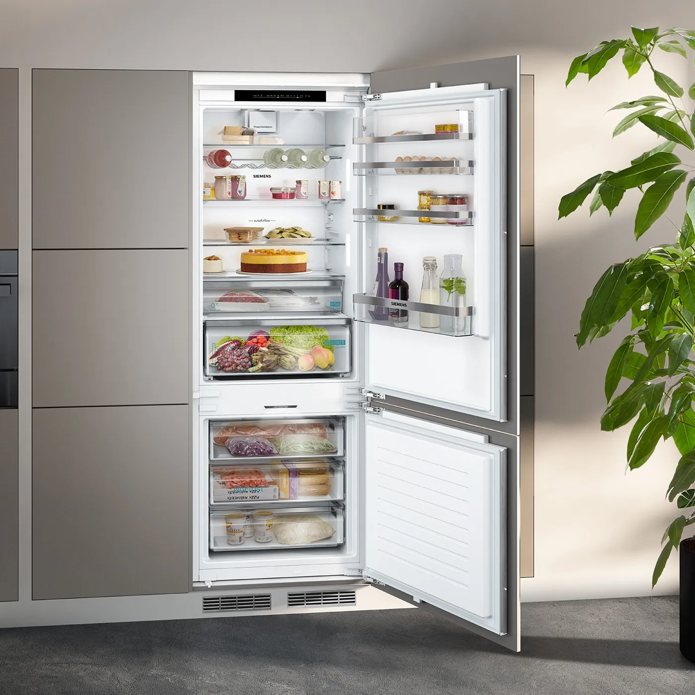 Et moderne køleskab i rustfrit stål med åbent dør, der afslører organiserede hylder fyldt med mad og drikkevarer, samt en fryser nedenunder med frosne varer. En grøn plante er synlig i forgrunden.