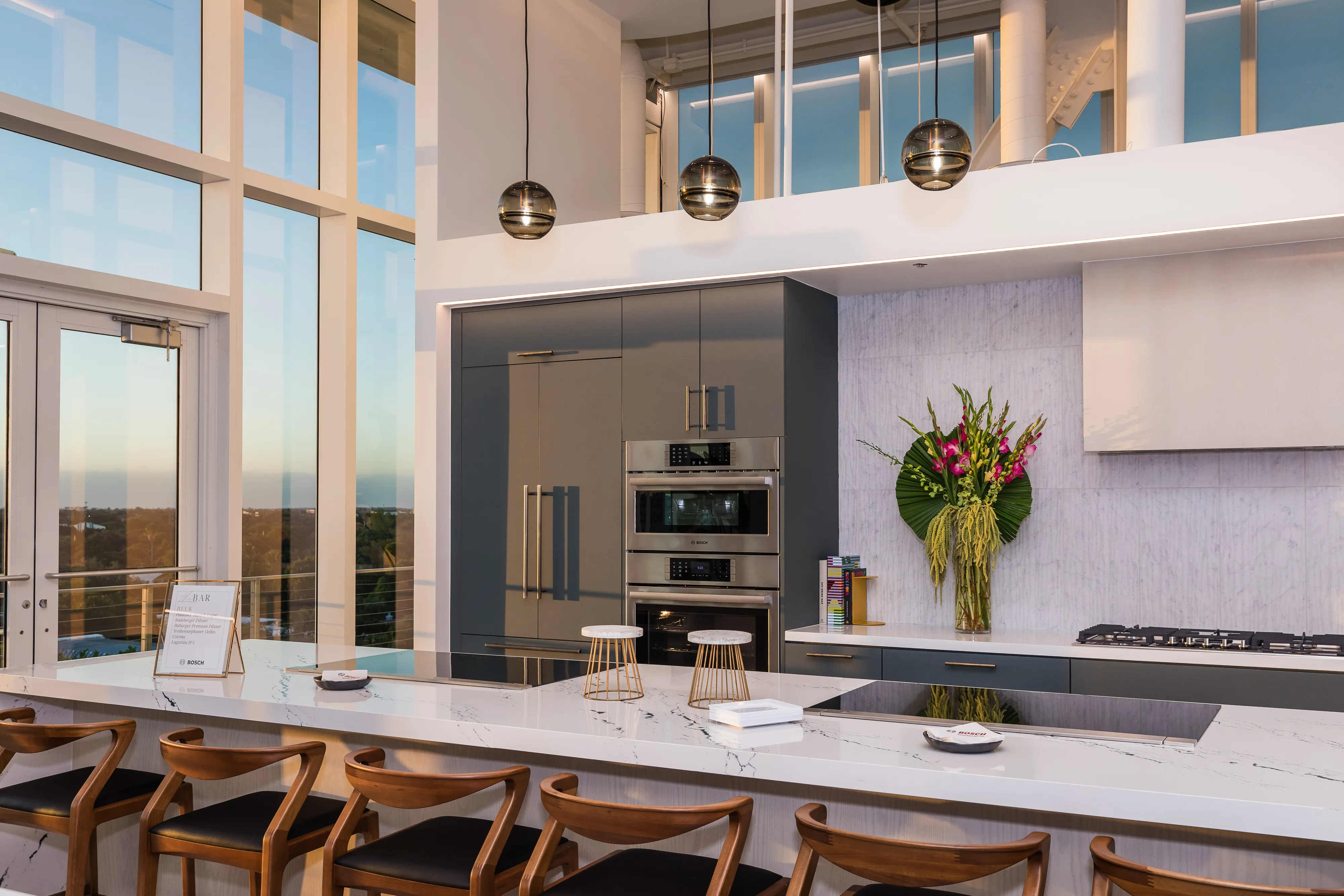 Modern kitchen with marble island, built-in Bosch ovens, and large windows. Pendant lights hang above.