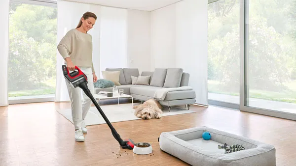 A woman uses a Bosch cordless vacuum cleaner to clean up spilled pet food on a wooden floor, with a dog and a dog bed in the background of a bright living room.