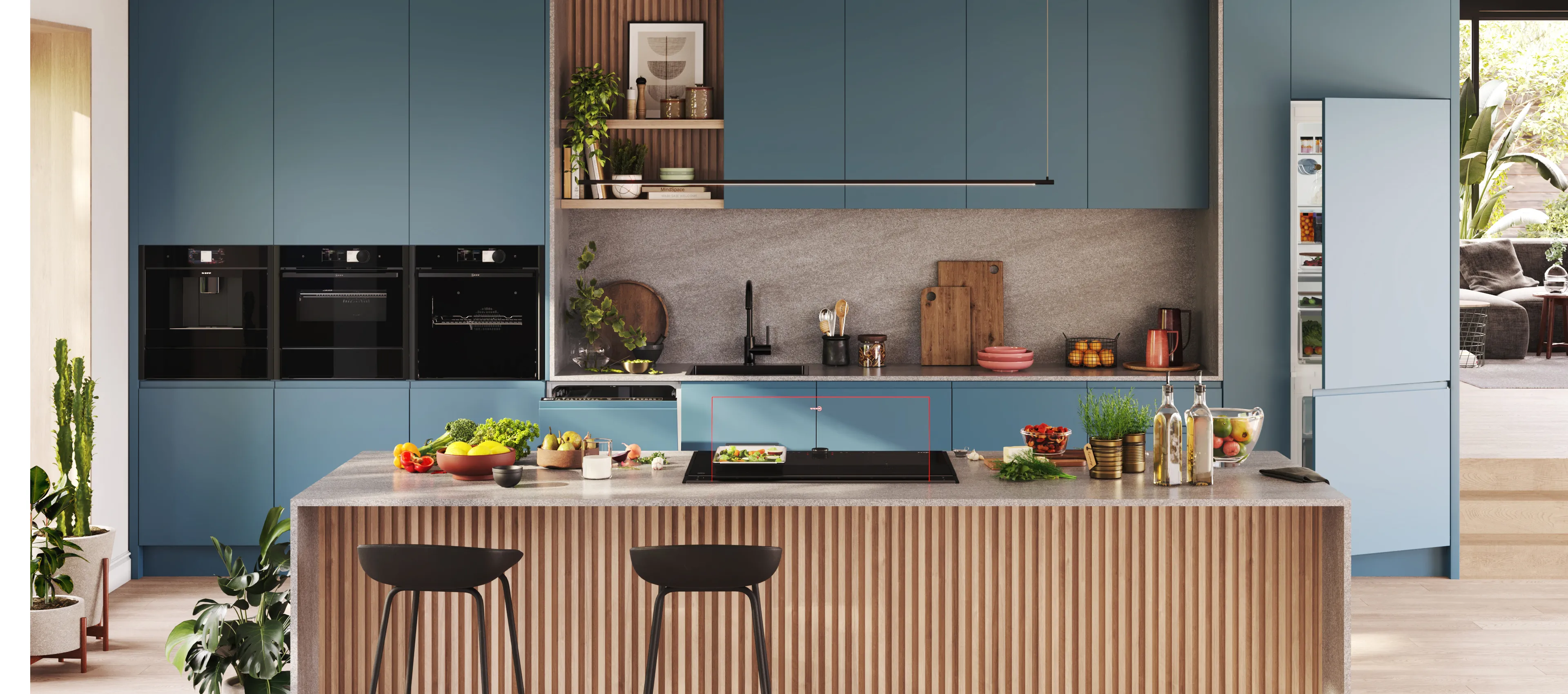 Modern open-plan kitchen with matte blue cabinets, wood‑slatted island and three black bar stools, stone countertop with bowls of fruit, cutting boards, plants and glassware, bright window.