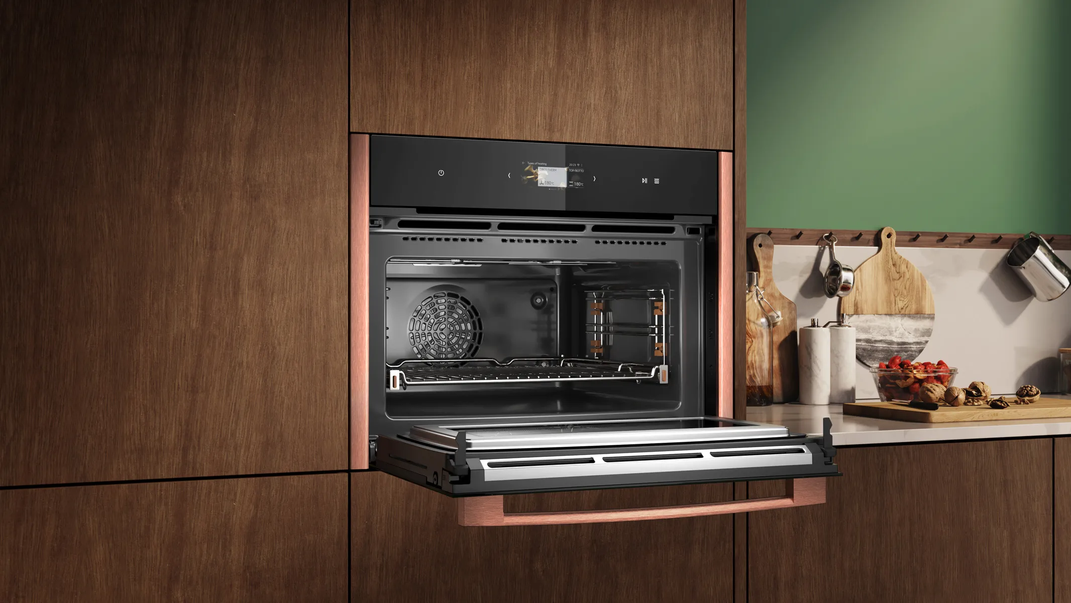 Built-in black-and-steel oven set into dark wood cabinets with the door open and the stainless-steel interior, fan and oven rack visible; to the right is a countertop with wooden cutting boards, jars, hanging utensils and a bowl of tomatoes against a green backsplash.