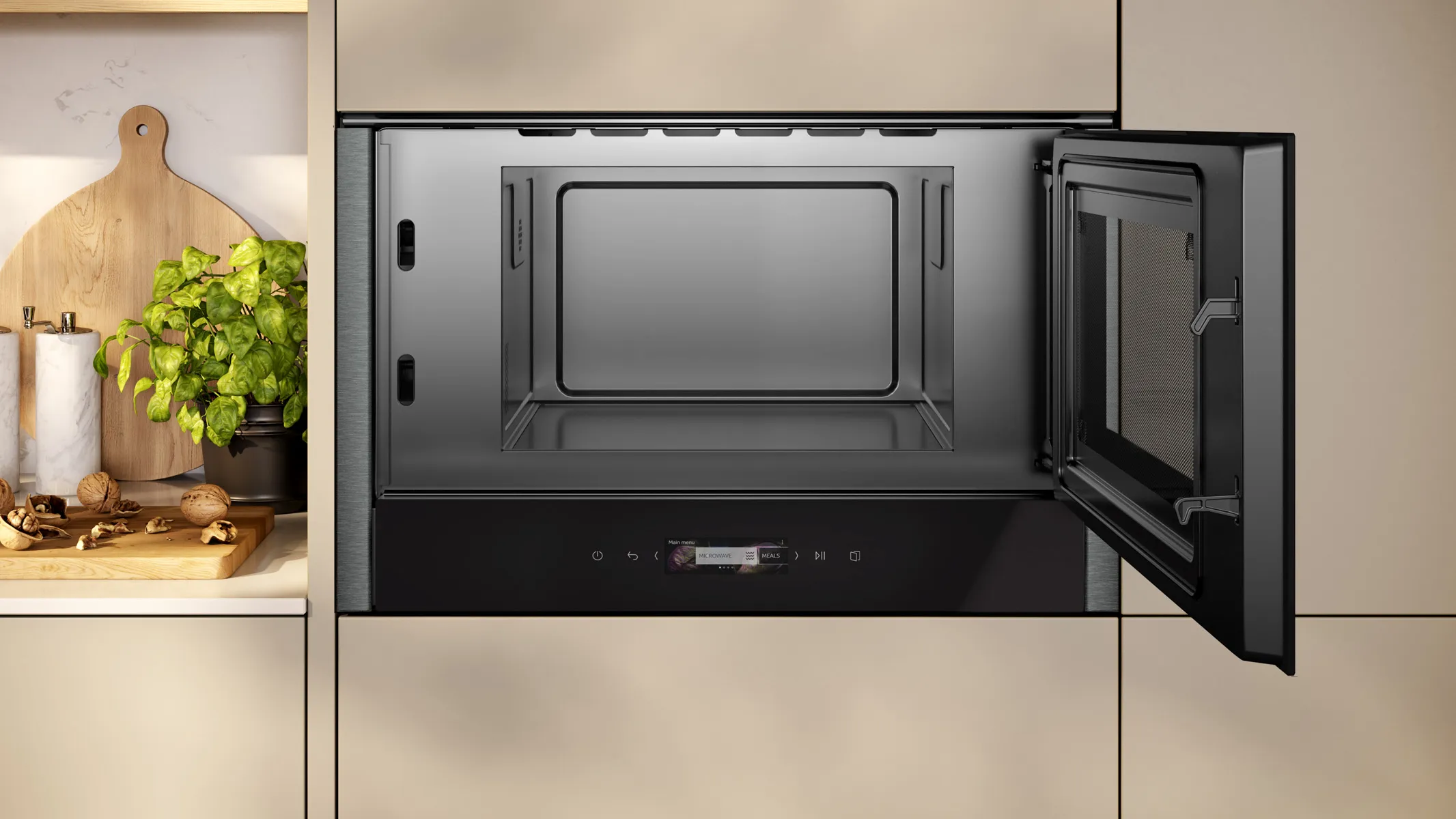 Built-in stainless-steel microwave with its door open inside beige kitchen cabinetry, empty interior visible; to the left on the countertop a wooden cutting board, a potted herb plant and small bowls.