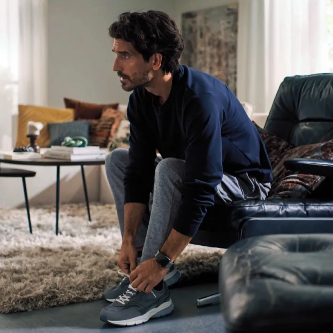 The image shows a man sitting on a couch, tying his shoes in a casual, indoor setting.
