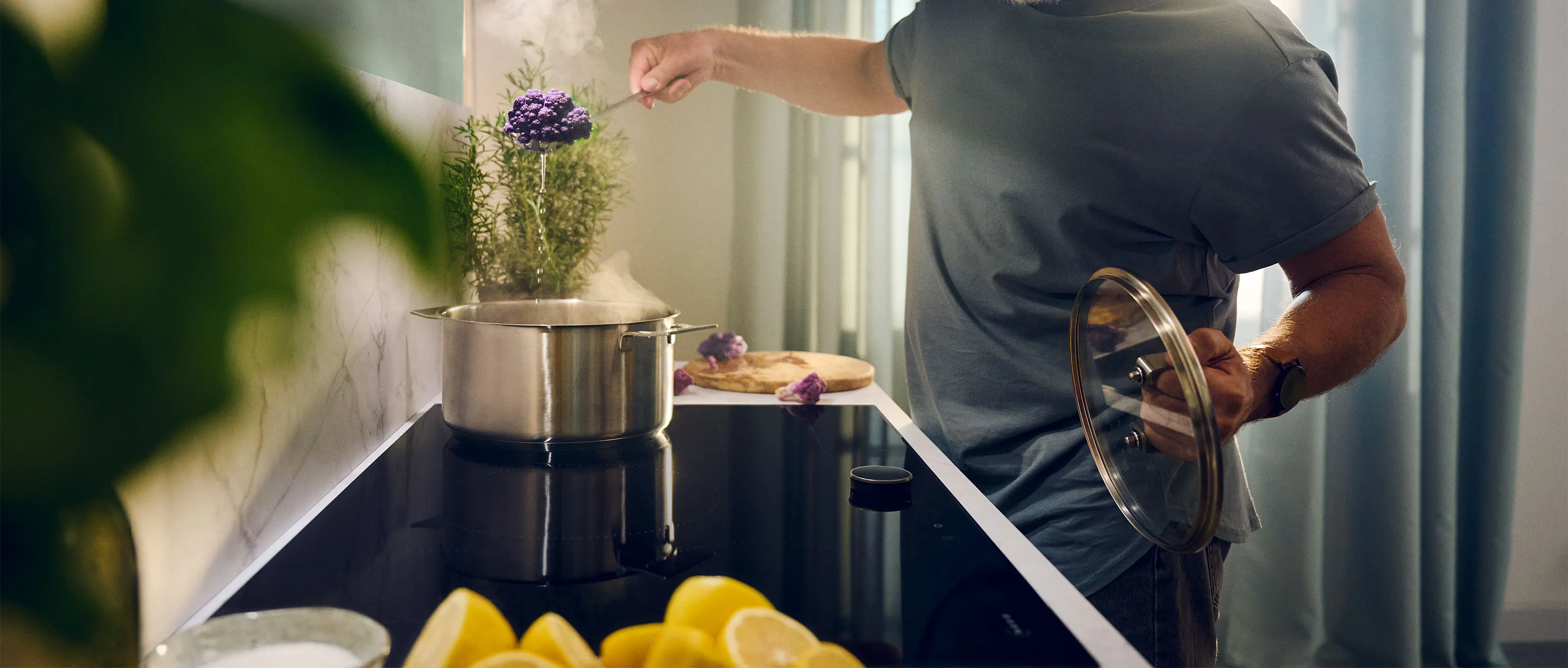 Cropped view of a person from the shoulders to waist in a home kitchen, wearing a gray shirt, lifting a stainless-steel pot lid with one hand and sprinkling seasoning into a steaming pot with the other. A small potted plant with purple flowers sits on the counter beside the stove and a wooden cutting board with chopped ingredients is visible behind the pot.