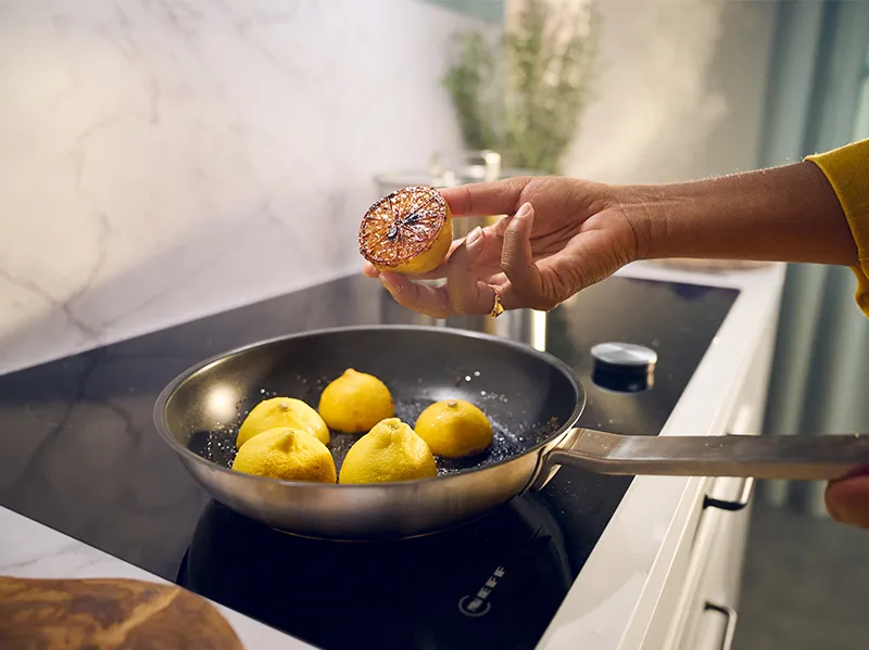 Hand in a yellow sleeve holding a seared lemon half over a frying pan of lemons on an induction cooktop.