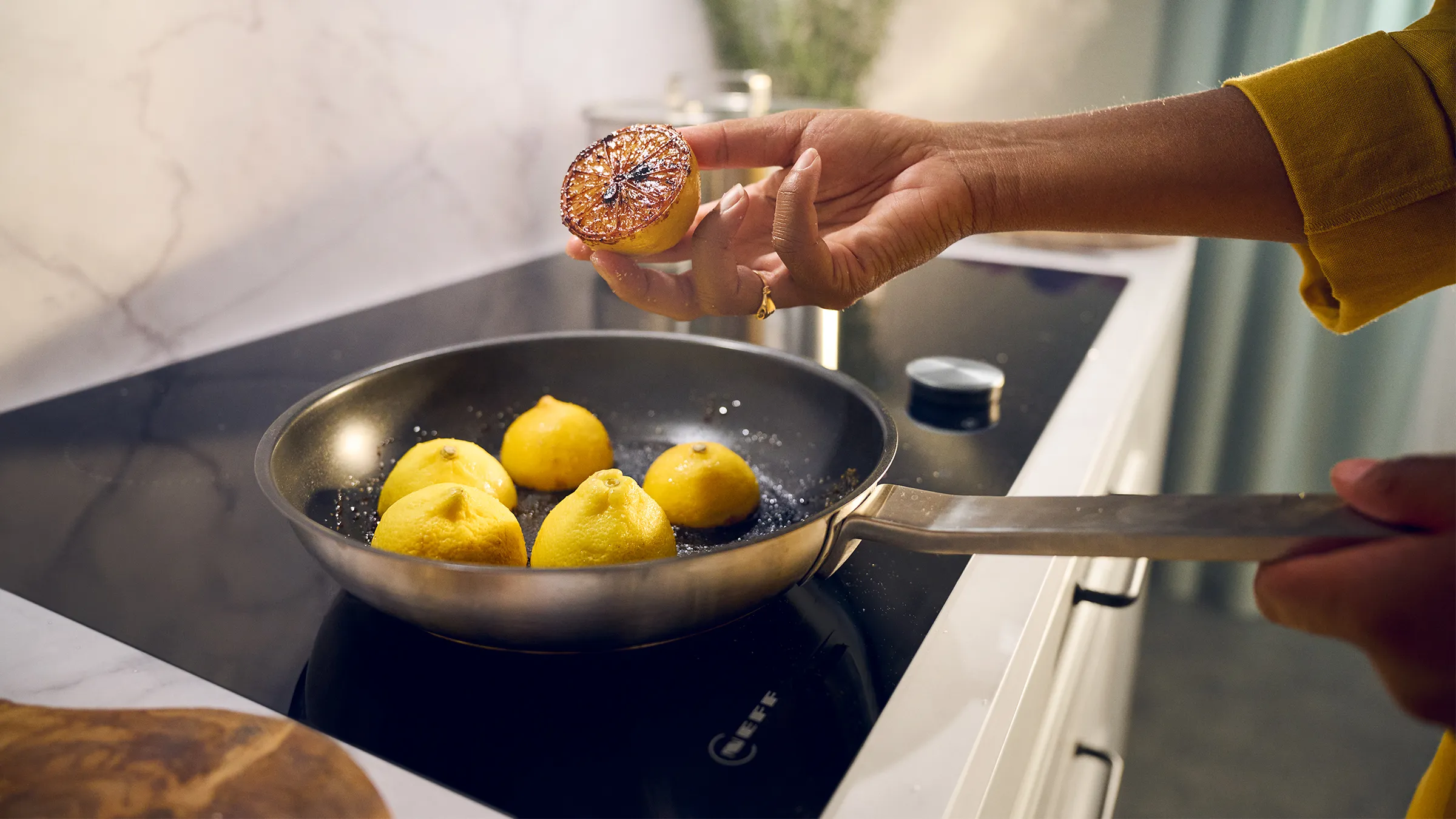 Hand in a yellow sleeve holding a seared lemon half above a frying pan of lemons on an induction cooktop.