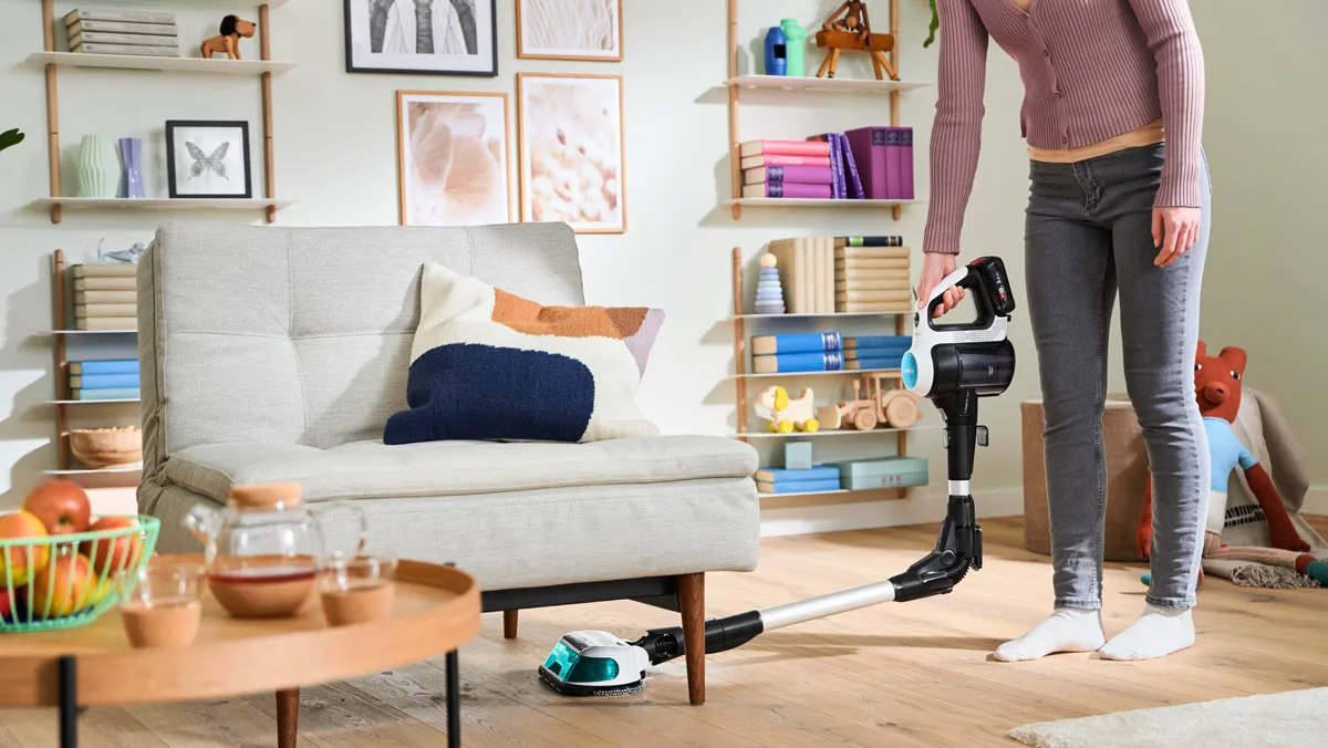 Person using a Cordless vacuum cleaner with hinged extension to clean under a sofa in a living room.