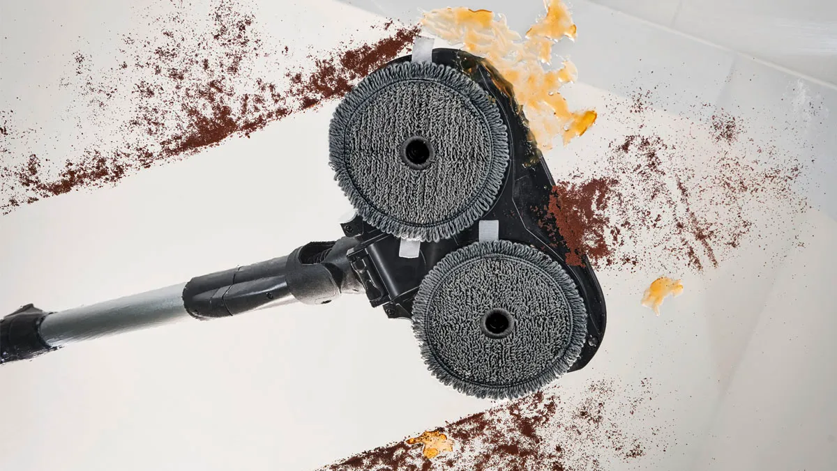 Underside of a vacuum head with two circular cleaning pads removing crumbs and food residue from a white floor.