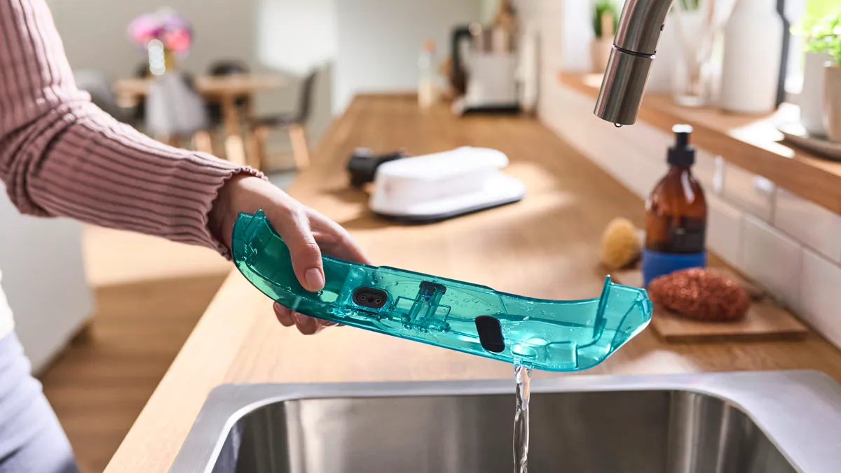 Hand holding a translucent teal water reservoir being rinsed under a kitchen tap.