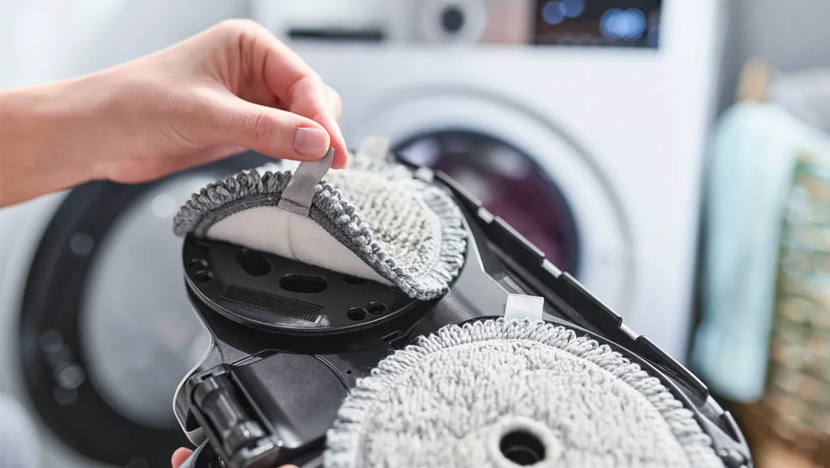 Hand lifting a microfiber pad from a dual-rotary cleaning head with a washing machine visible in the background.