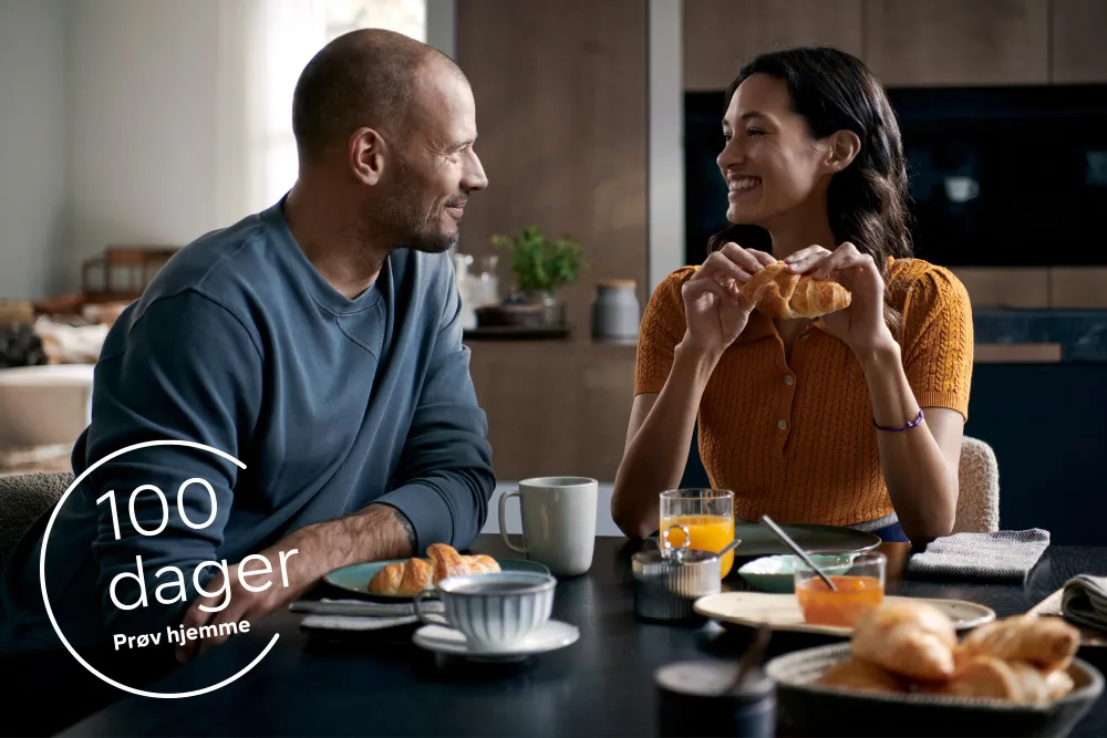 Et par nyter frokost sammen ved et koselig kjøkkenbord med croissanter, kaffe og appelsinjuice i moderne omgivelser.