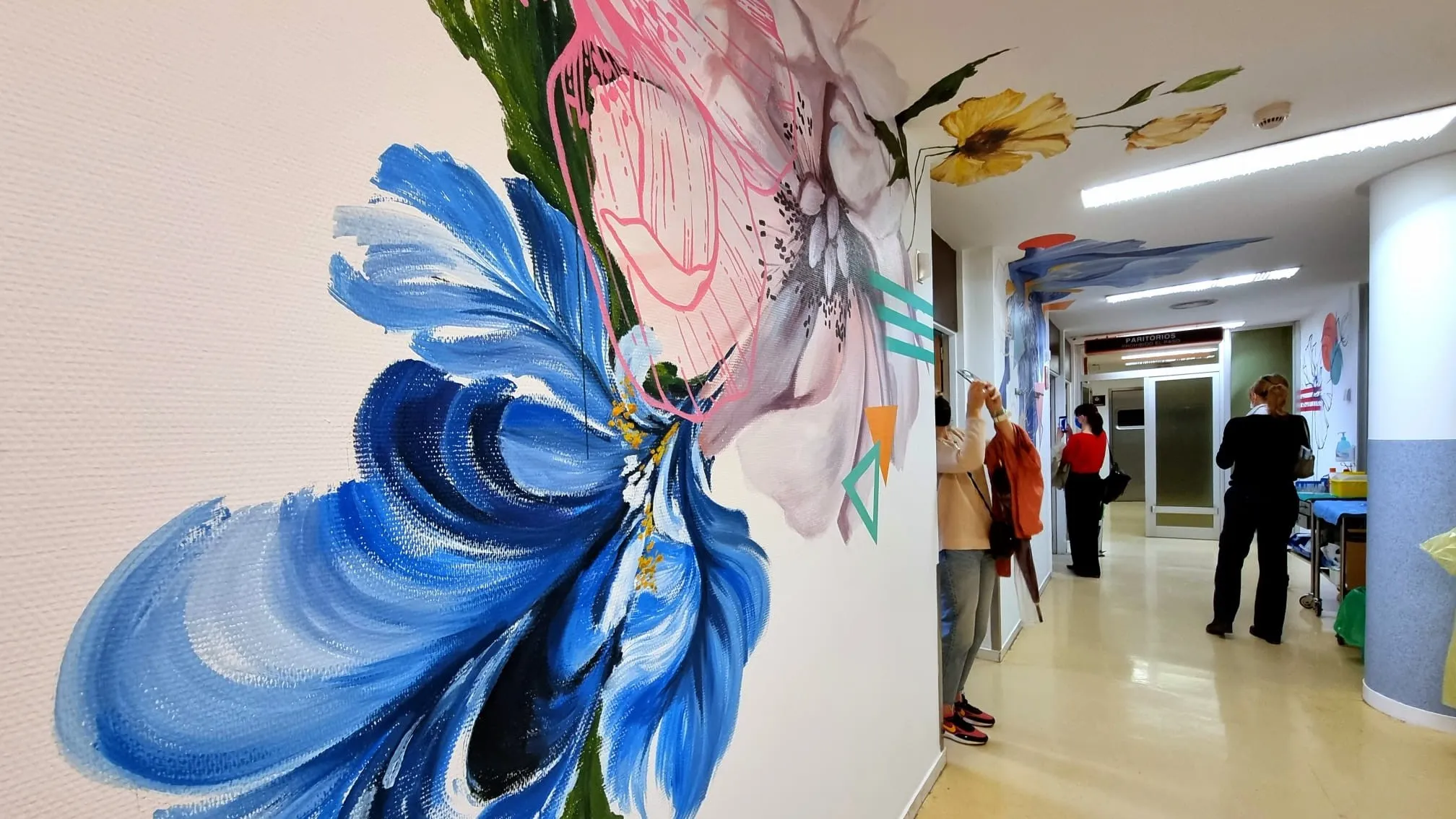 Mural colorido de flores en pasillo, con personas observando el arte en un espacio moderno y acogedor. 