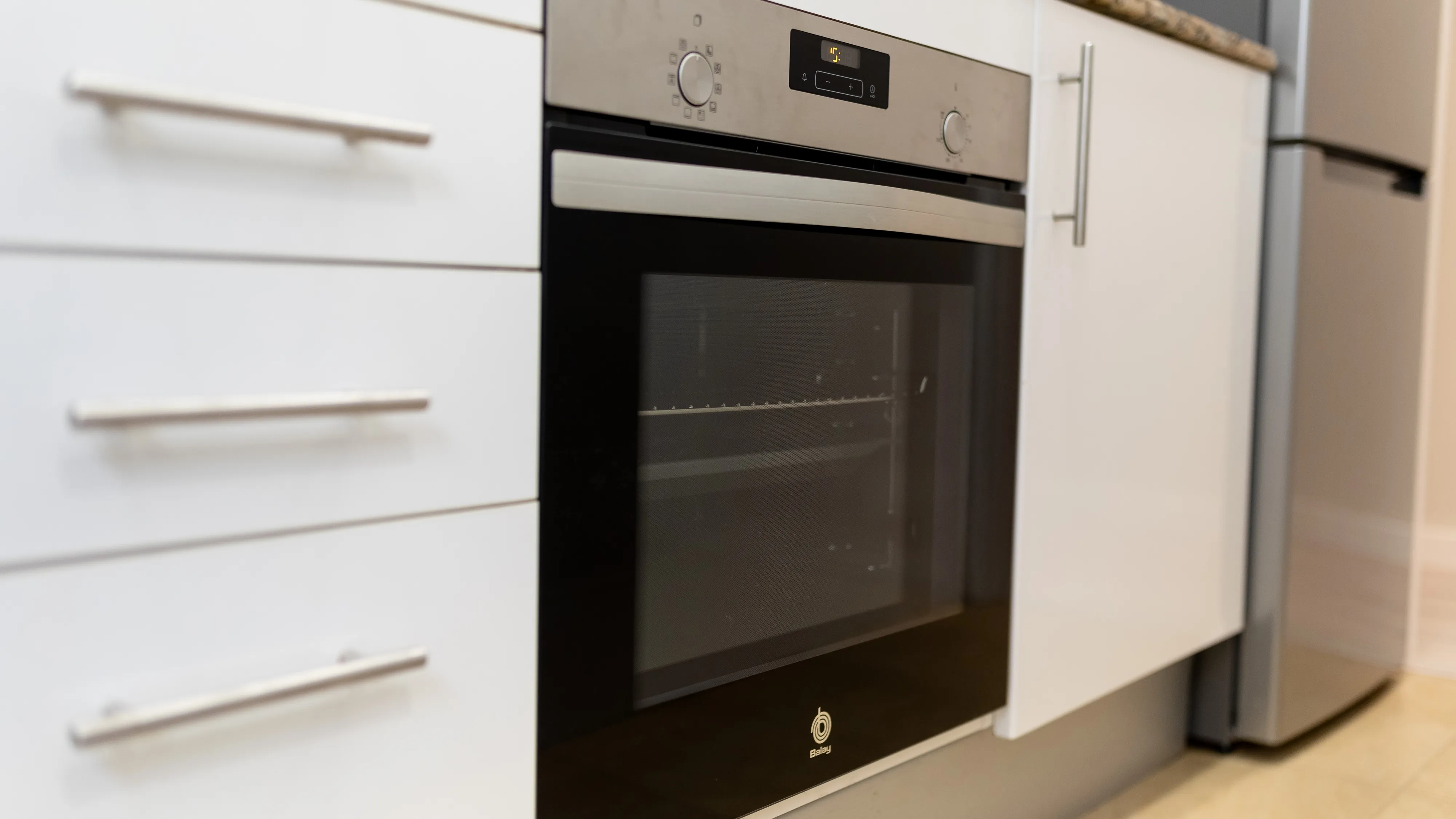 Horno de cocina moderno con controles digitales y puerta de vidrio, parte de una configuración de gabinete de cocina blanco. 