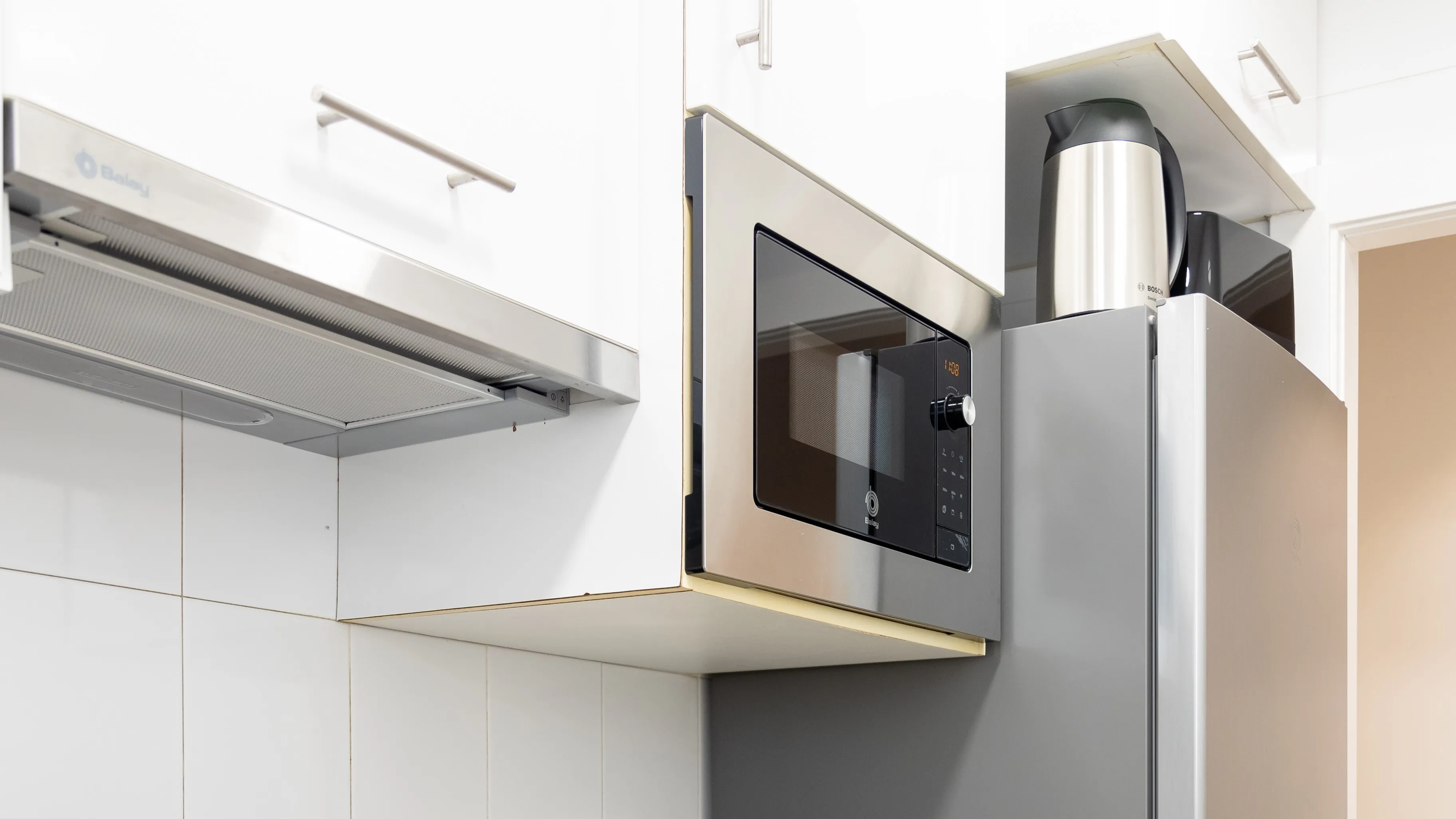 Cocina moderna con microondas de acero sobre refrigerador, gabinetes blancos y tetera al lado. 