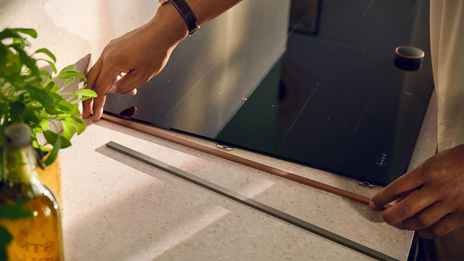 Hands measuring and cutting wooden plank on kitchen counter with potted plant.