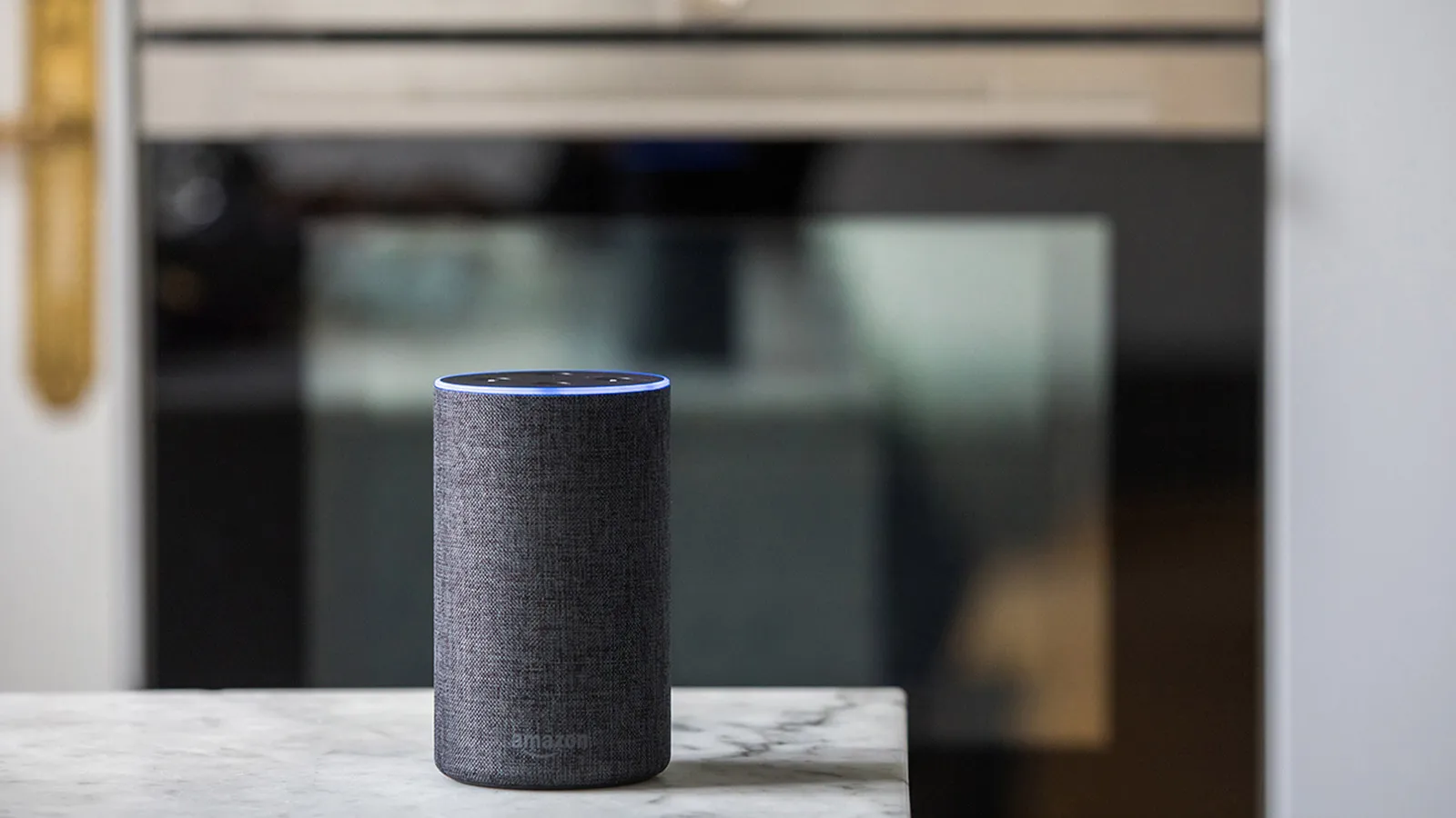 An Amazon Echo smart speaker, with its blue light activated, sits on a white marble kitchen countertop. In the background, a modern built-in oven is out of focus.