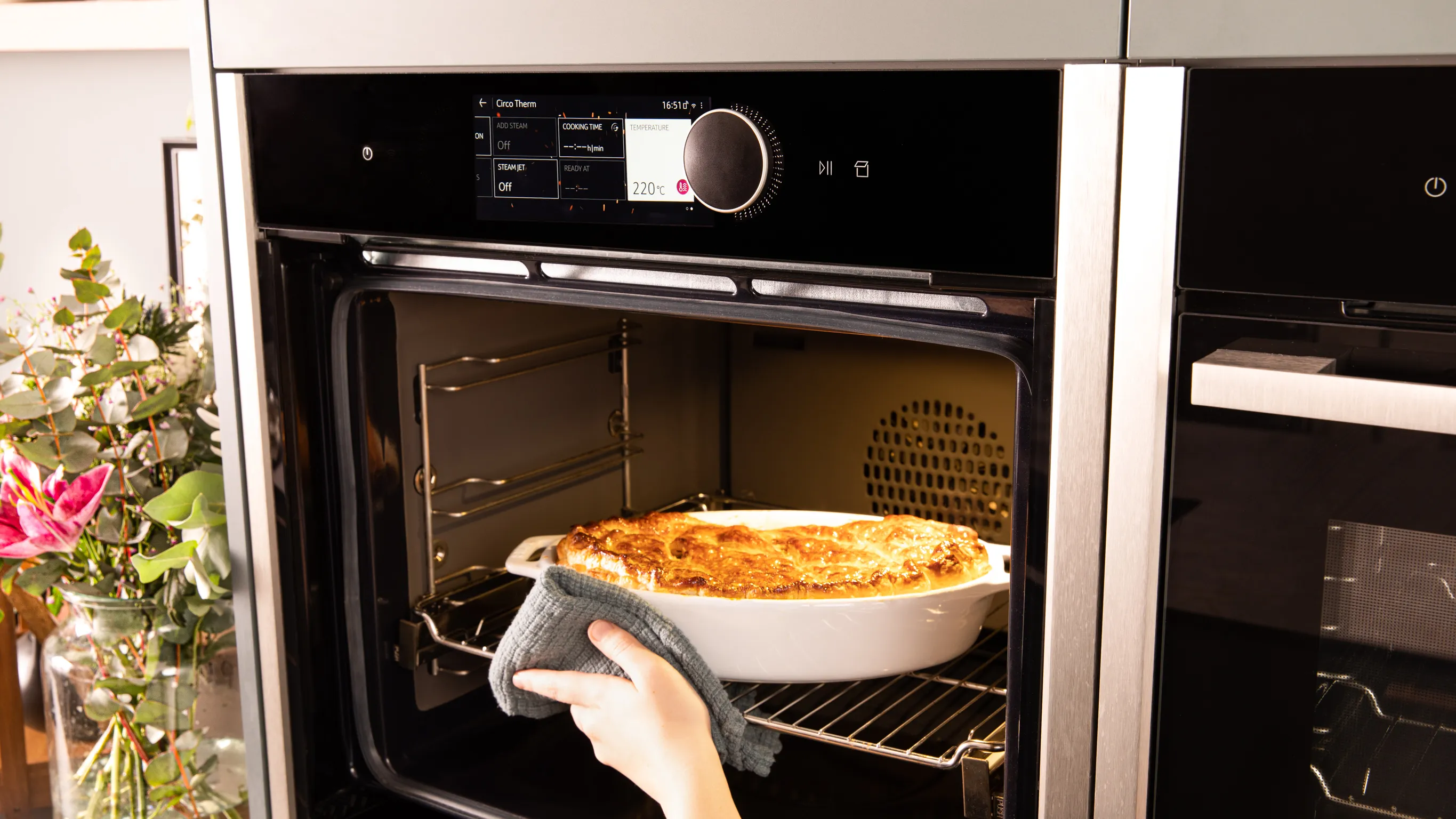 Hand nimmt eine Auflaufform mit überbackenem Gericht mithilfe eines Topflappens aus einem modernen Einbaubackofen mit digitalem Display und Teleskopauszug.