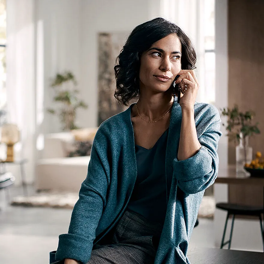 Das Bild zeigt eine Frau mit dunklen lockigen Haaren, die einen teal Cardigan tragen, sitzen und nachdenklich aussehen.