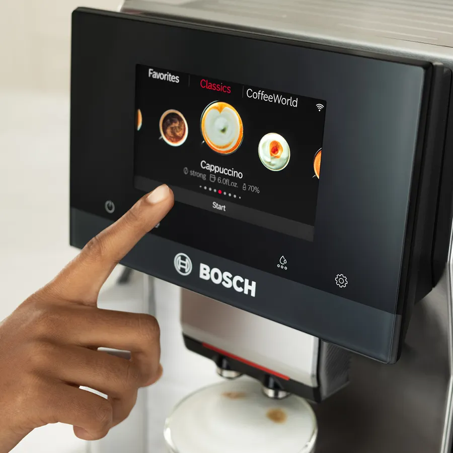 Finger selecting cappuccino on a Bosch coffee machine touch screen interface.