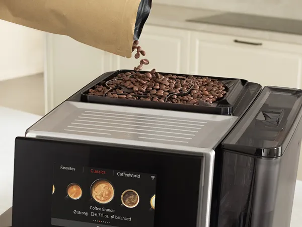Coffee beans being poured into the top compartment of a Bosch coffee machine with a digital display.
