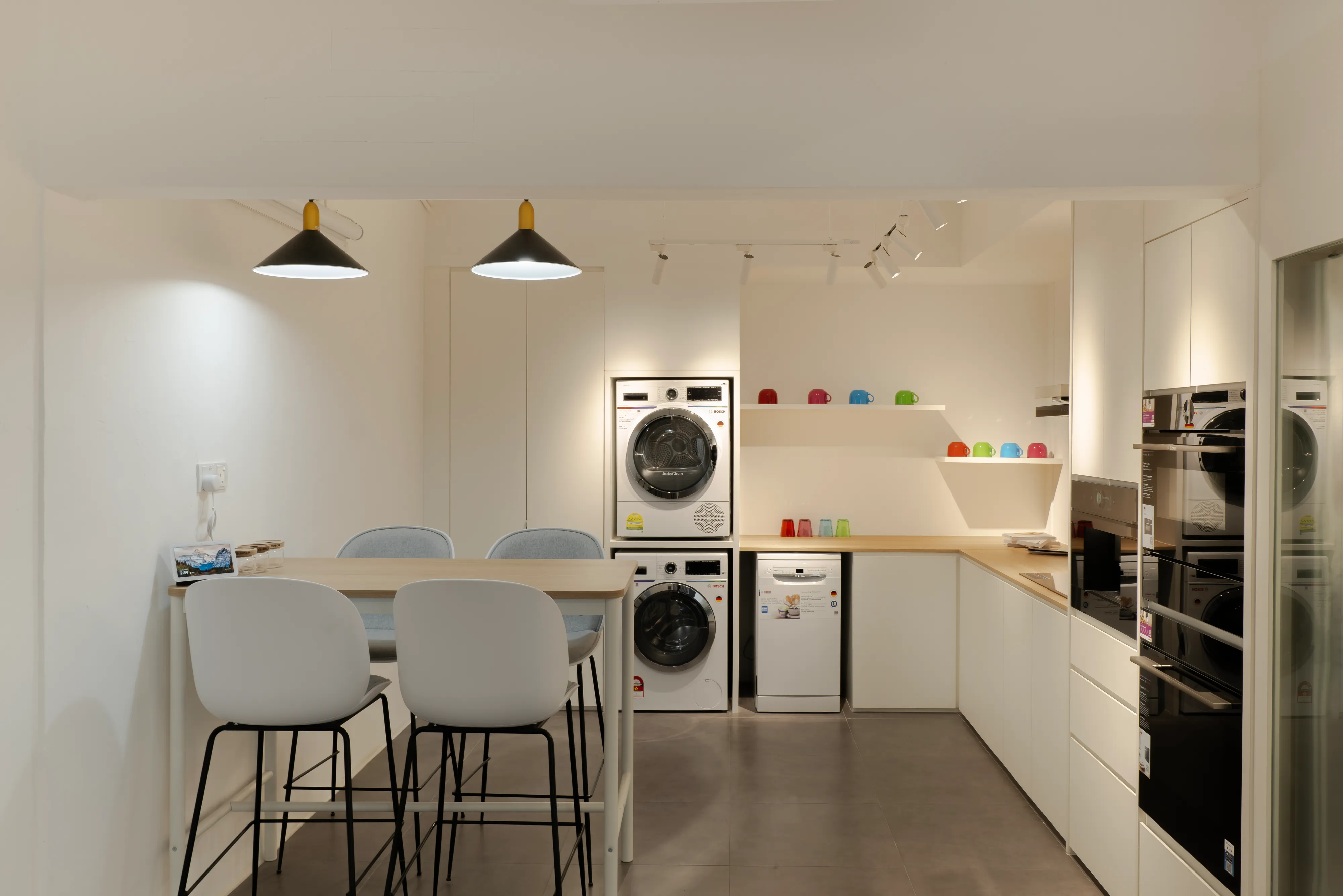 Bosch showroom kitchen and laundry concept area with high table seating, stacked washer-dryer units, freestanding dishwasher, and built-in ovens in modern white cabinetry.