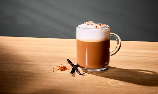 Vanilkové Latte Macchiato na drevenom stole s kúskami škorice vedľa neho.