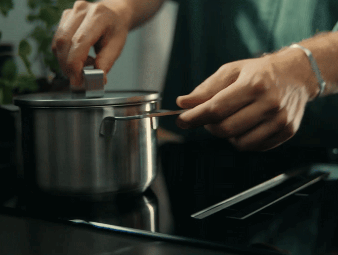 Hands stirring a pot on a stove, cooking utensils, kitchen scene.
