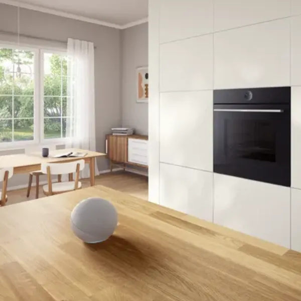 A modern kitchen featuring a black built-in oven Bosch, a light wood countertop with a spherical smart speaker, and a dining area with a window in the back.