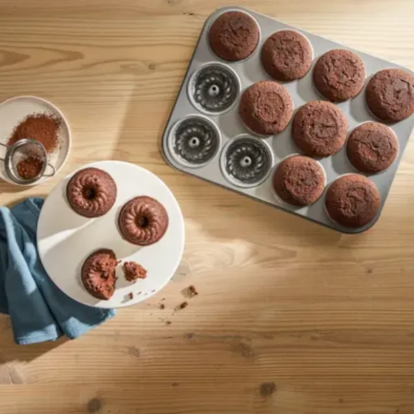 Un vassoio con mini bundt cake al cioccolato appena sfornate, tre tortine su un piatto e cacao in polvere su un tavolo di legno.