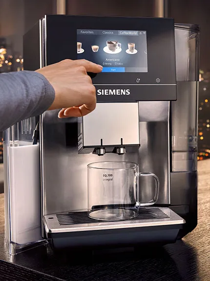Dłoń obsługuje ekspres Siemens, wybierając Americano na dotykowym ekranie, obok przezroczystej filiżanki
