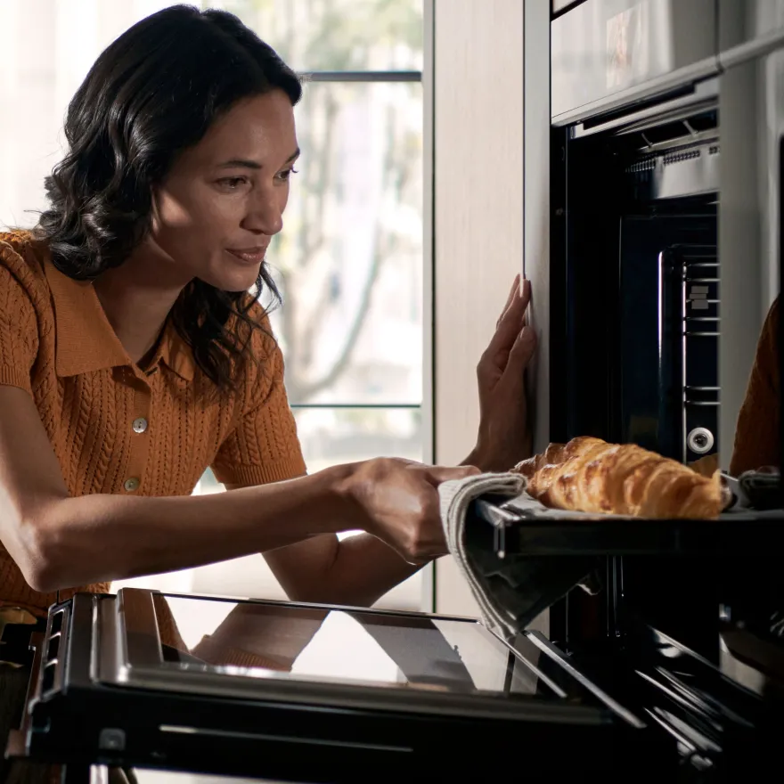 A woman with dark, wavy hair, wearing an orange knit top, is carefully taking a freshly baked croissant out of a modern, black built-in oven. She is using a cloth to hold the hot baking tray.