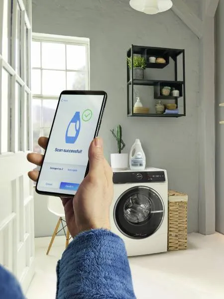 Smartphone in de hand met het bericht "Scan succesvol", wasmachine en kast in een interieur.