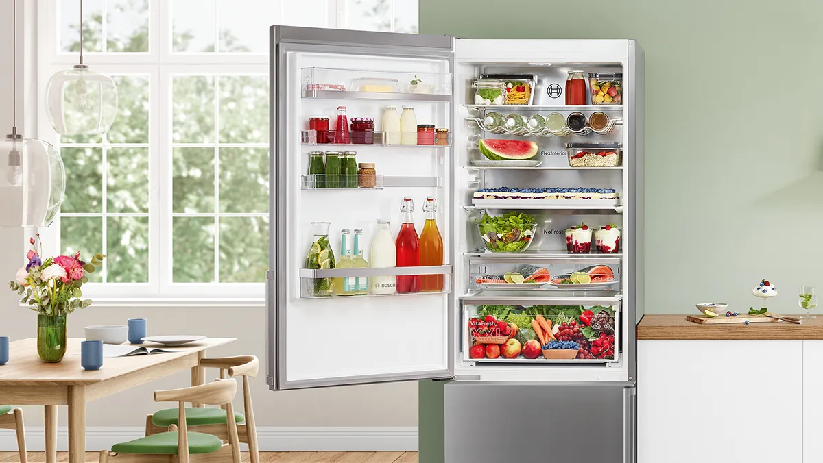 A Bosch freestanding fridge-freezer, its door open to reveal colourful fresh food, situated in a bright, modern kitchen with natural light.