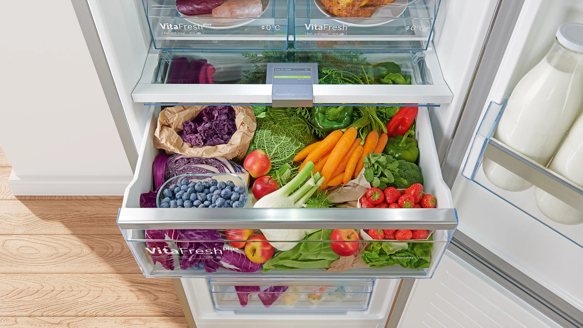 Crisper drawer inside a fridge filled with fresh vegetables, fruits, and leafy greens including carrots, strawberries, blueberries, and lettuce.