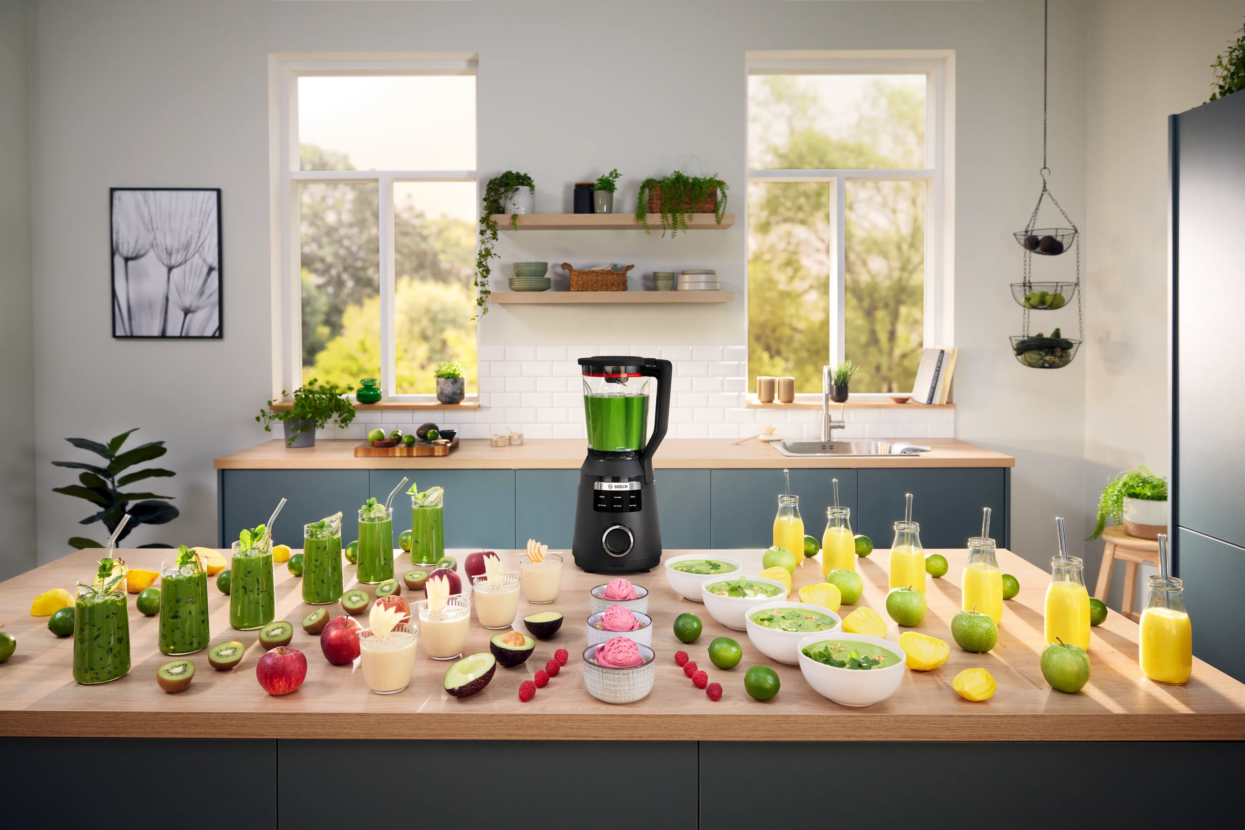Modern kitchen with blender on island surrounded by various fresh fruits, vegetables, and smoothies in glasses and jars.