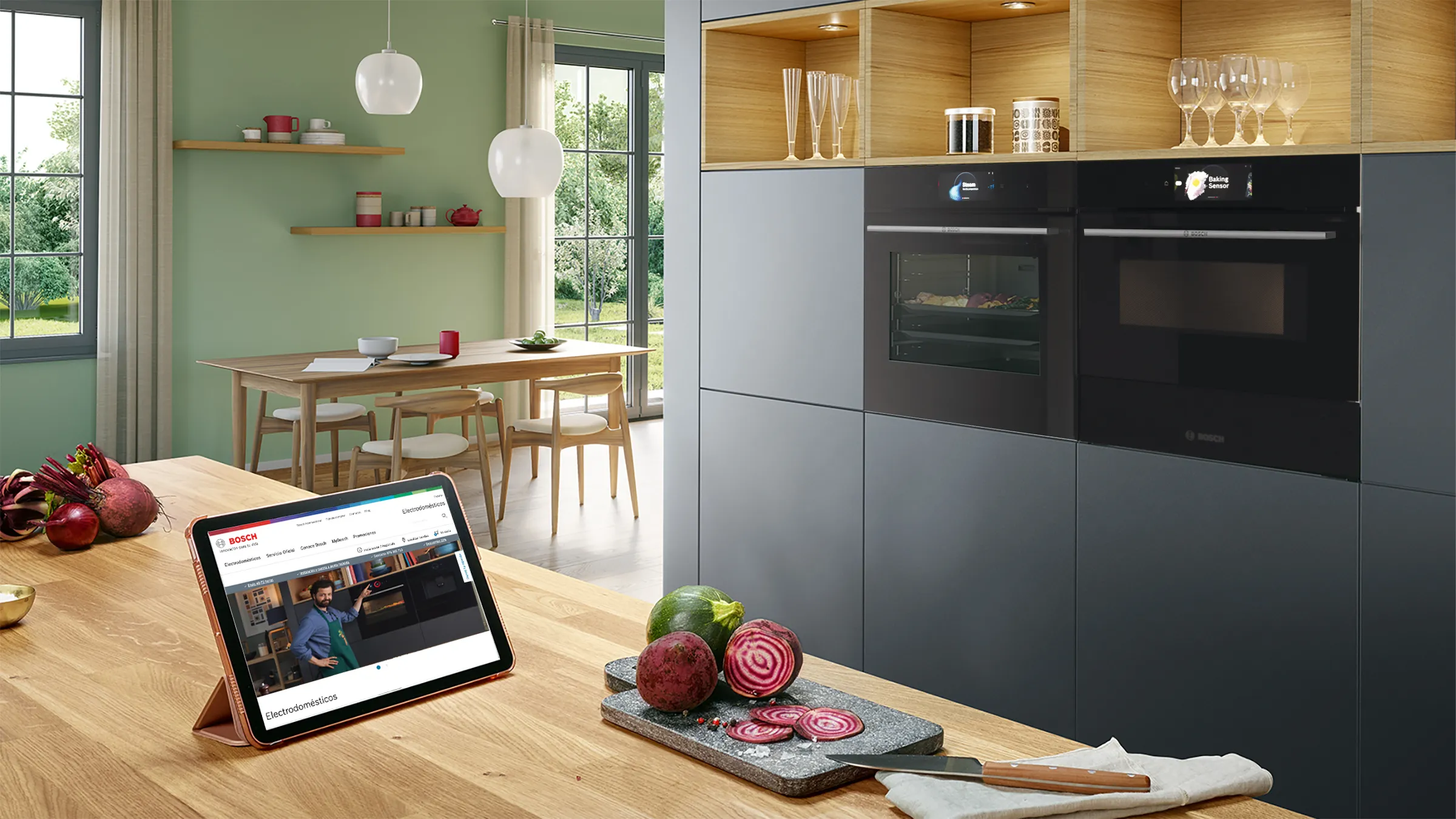 Cocina moderna con armarios de madera, electrodomésticos, mesa de comedor y tabletas mostrando vídeo de cocina.
