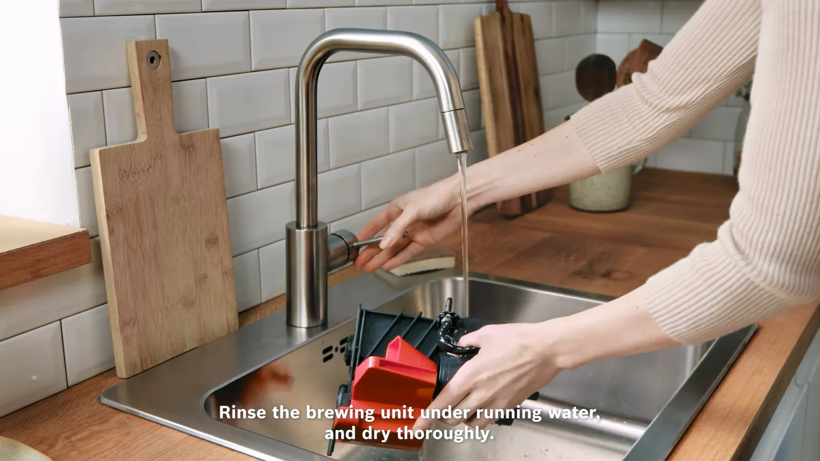 Person rinsing Bosch coffee machine brewing unit under a kitchen faucet with tiled backsplash.