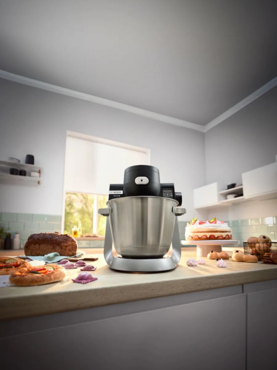 En elegant svart köksmixer på en bänkskiva, omgiven av olika bakade varor, inklusive pizza, tårta och kakor, i ett ljust kök.