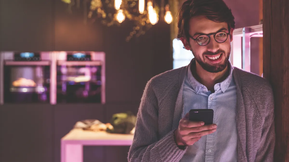 Un uomo sorridente con in mano uno smartphone appoggiato alla porta di una cucina illuminata con luci da atmosfera