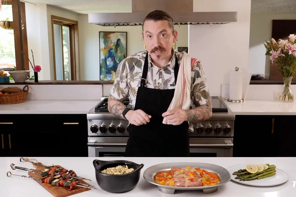 Man wearing a patterned shirt and black apron standing behind a kitchen counter with prepared food dishes including grilled vegetables, a pot of pasta, a pan of cooked food, and sliced cucumbers