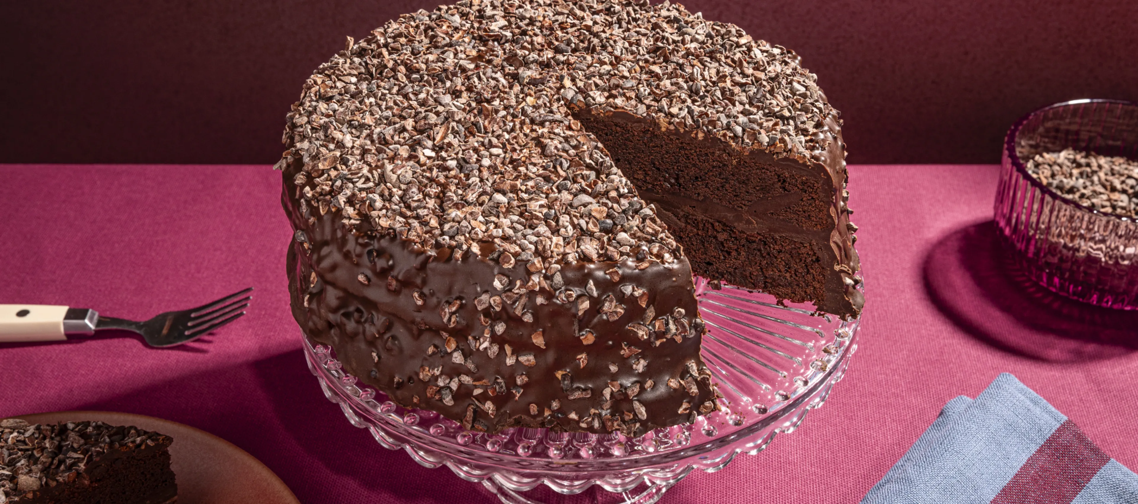 Chocolate cake with nuts and chocolate frosting on a glass cake stand against a burgundy background.
