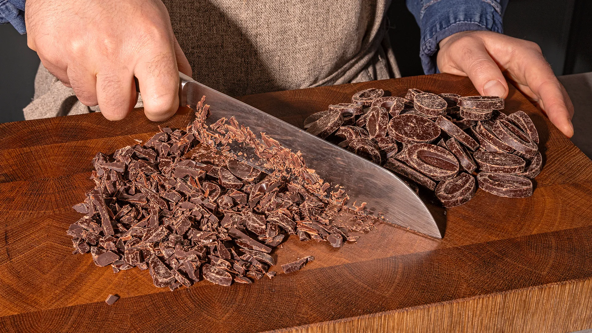 Hands chopping dark chocolate on a wooden cutting board.