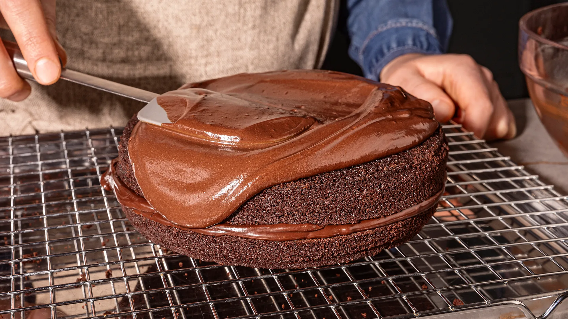 Chocolate cake being frosted on a cooling rack, hands applying chocolate glaze.