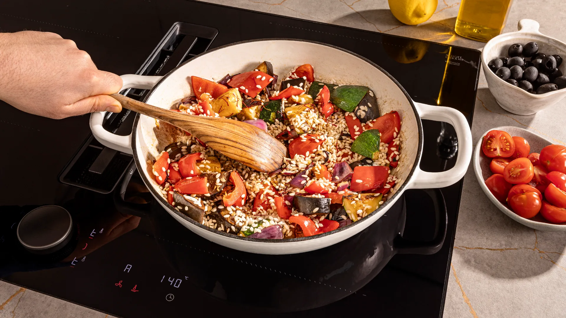 Cooking pan with healthy vegetable stir-fry, tomatoes, olives, and cooking utensils on stovetop.