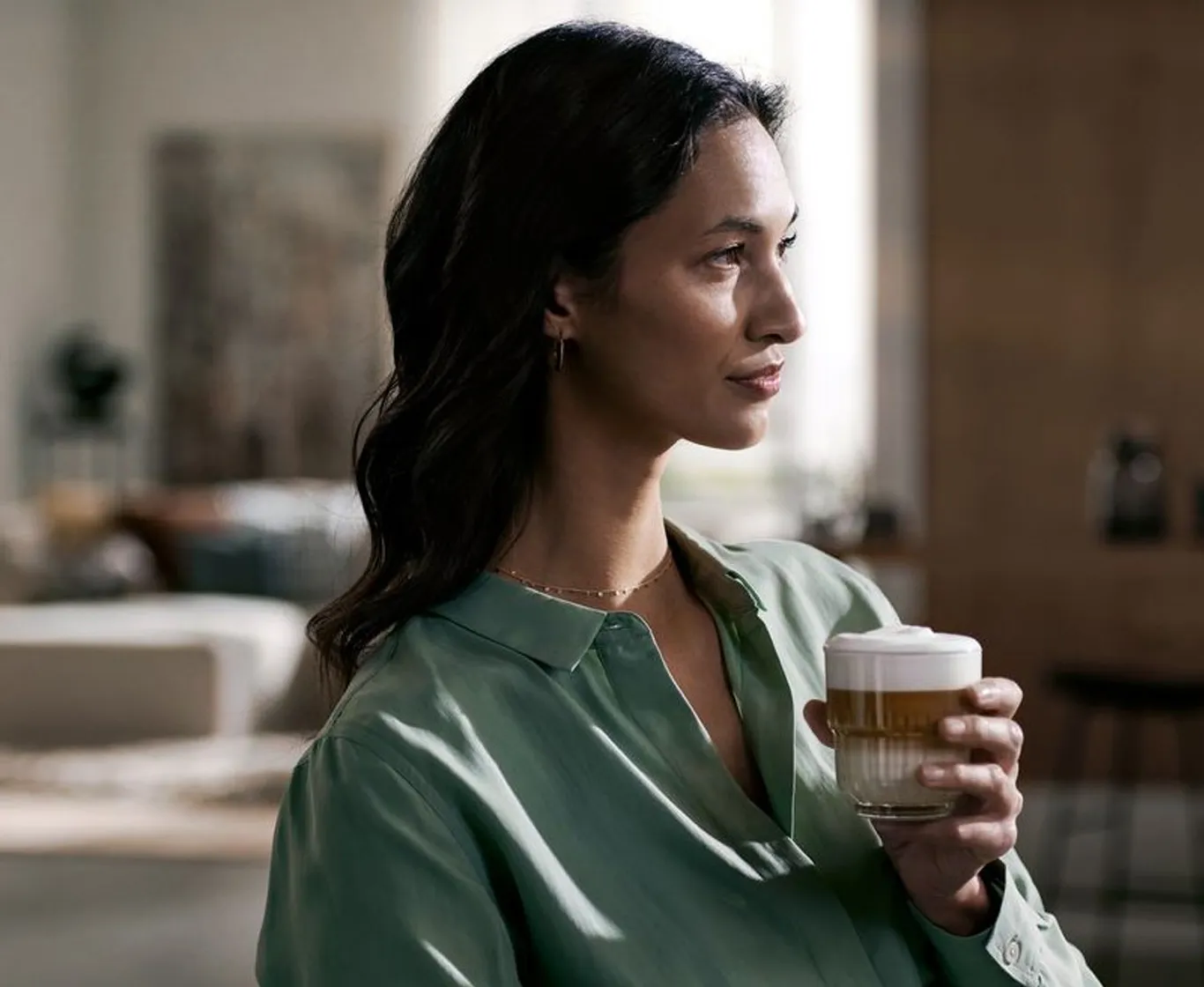 Eine Frau hält ein Glas Latte Macchiato und blickt nachdenklich zur Seite.