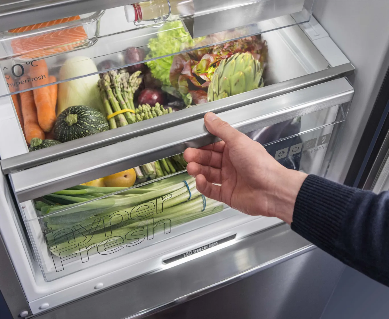 Eine Hand öffnet eine hyperFresh Kühlschrank-Schublade voller frischem Gemüse und Lachs.